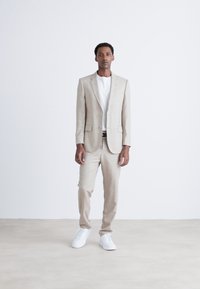 Zweiteiliges Anzugset aus leichtem beigefarbenem Leinen, kombiniert mit einem weißen T-Shirt und weißen Sneakers. Minimalistisches Design mit strukturiertem Blazer und schmal geschnittenen Hosen.