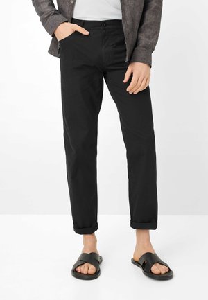 Persona che indossa pantaloni neri risvoltati, sandali neri con cinturini incrociati, una camicia bianca e una giacca grigio scuro, in piedi con una mano in tasca.