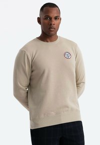 Sudadera beige con cuello redondo, mangas largas y un parche circular con el logo en el lado izquierdo del pecho. Tejido suave y texturizado.