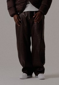 Isik kannab tumeda pruuni nahkpükse, valgeid spordijalatsi, tumeda pruuni puffjakki ja valget särk, seistes ühetaolisel taustal.