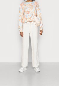 Persona con camisa de manga larga floral, pantalones de pierna ancha color blanco roto, zapatillas blancas y un bolso cruzado beige contra un fondo liso.