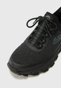 Zapatilla deportiva negra con parte superior de malla, cordones texturizados y puntera reforzada. Presenta una suela gruesa con patrones de tracción pronunciados.