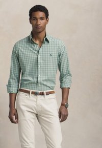 Camicia button-down a quadretti verdi e bianchi, maniche lunghe arrotolate, abbinata a pantaloni beige e cintura marrone. Logo polo ben visibile sul petto.