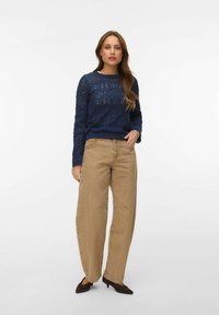 Femme portant un pull en tricot à motifs bleu marine, un jean large couleur sable et des escarpins sombres à bout pointu, debout devant un fond blanc.