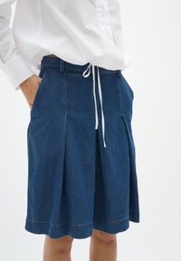 Denim krilo v temno modri barvi s gubami, pasom na vrvico in stranskimi žepi. Ima kontrastno šivanje ob robu. Gladka tekstura.