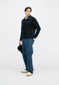 Fiatal férfi texturált sötétkék és fekete pulóverben, laza kék farmernadrágban, fehér tornacipőben, egy fekete sapkát tartva, sima háttér előtt áll.