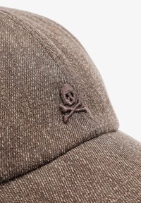 Gorra marrón con una tela texturizada y un logo de calavera y huesos cruzados bordado en marrón oscuro en el panel frontal.