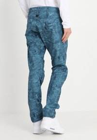 Blue patterned cargo pants with a relaxed fit, featuring multiple pockets and a smooth, textured fabric. White sneakers complete the look.