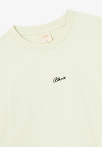 T-shirt en coton vert clair avec un col rond, arborant un petit logo noir brodé sur la poitrine et une texture douce.