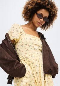 Γυναίκα με σγουρά μαλλιά που φοράει μικρά μαύρα γυαλιά ηλίου, κίτρινο φόρεμα με λουλούδια και καφέ υπερμεγέθη σακάκι περασμένο στους ώμους.