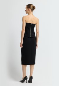 Černý top bez ramínek s přiléhavým střihem, se zřetelným zlatým zipem na zadní straně. V kombinaci s ladící tužkovou sukní a černými podpatky.