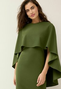 Grön klänning med en cape-liknande överdel med vågiga kanter, tillverkad av ett slätt tyg. Elegant design, skräddarsydd passform och korta ärmar.