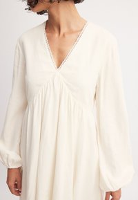 Langarm, jurk tot de knie in een off-white stof. Heeft een V-halslijn met delicate kantafwerking en geplooide details onder de buste.