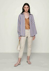 Ριγέ overshirt σε μπλε και άσπρο, μπεζ μπλούζα, κρεμ παντελόνι, λευκά σανδάλια, κρατώντας μία μικρή λευκή τσάντα.