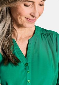 Blusa verde con una texture morbida e liscia, caratterizzata da un colletto a V e bottoni frontali. Il tessuto è leggero con un leggero lucido.