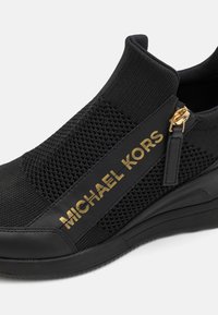 Baskets noires à enfiler avec dessus en maille, logo "Michael Kors" doré sur le côté, et détail de fermeture éclair dorée près de la cheville.