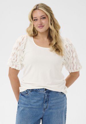 Blonde vrouw met golvend haar draagt een wit topje met gestructureerde pofmouwen en een blauwe spijkerbroek, staat met de handen achter de rug en glimlacht licht.