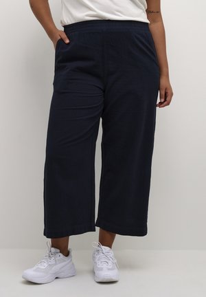 Persoon die zwarte wijde cropped broeken, witte sneakers en een wit shirt draagt met ontspannen handen langs de zij.