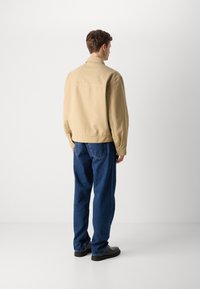 Beige bomulljakke med avslappet passform, synlige sømmer og krage; parret med løse blå denimjeans og svarte sko.