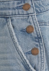 Vaaleansiniset farkushortsit, joissa on kolme ruskeaa nappia, joissa on kohokuvioitua tekstiä. Kankaassa on kulunut reuna ja tekstuuri, joka on karu.