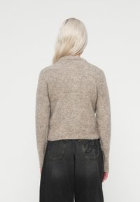 Μπεζ, μαλακό πλεκτό πουλόβερ με μακριά μανίκια και στρογγυλή λαιμόκοψη, φορεμένο πάνω από σκούρες τζιν παντελόνες με πίσω τσέπες και λεπτομέρειες κεν�τήματος.