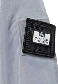 Gray fabric with a smooth texture features a black rectangular patch labeled "WEEKEND OFFENDER" with a crown emblem. Detailed stitching visible.