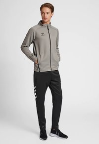 Grijze jas met ritssluiting en zwarte accenten, gecombineerd met zwarte broek met witte strepen. Het materiaal lijkt zacht en sportief.