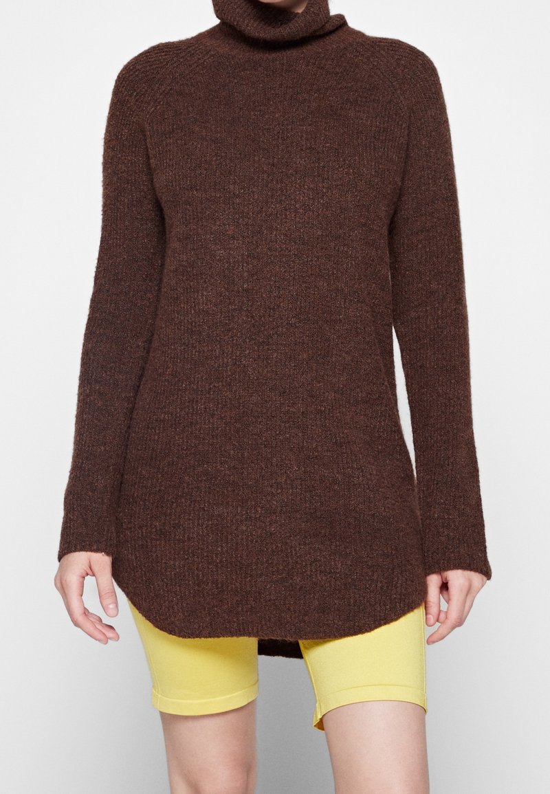 Pull en laine marron à manches longues et col roulé avec un tricot texturé, présentant un col haut et un ourlet arrière plus long, porté sur un short jaune.