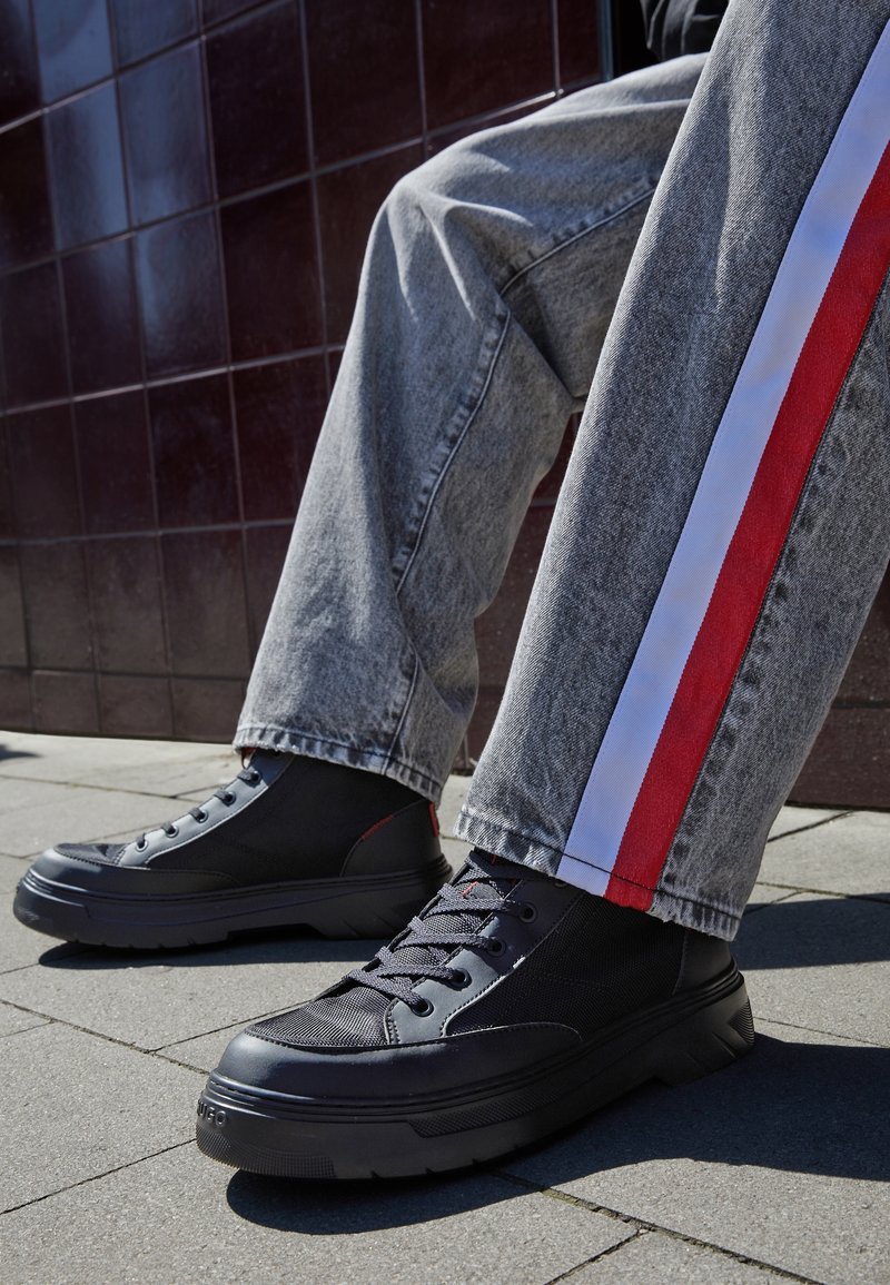 Personne portant des baskets montantes noires et un jean gris avec des bandes rouges et blanches sur les côtés, se tenant sur un pavé carrelé près d'un mur sombre.