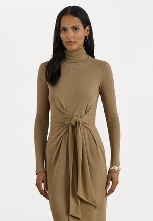 Woman with long dark hair wearing a beige long-sleeve turtleneck dress tied in a knot at the waist, accessorized with gold hoop earrings and bracelets.