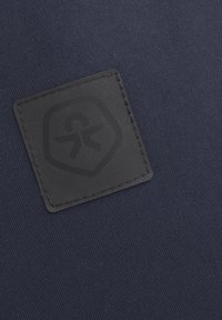 Mörkblått tyg med en strukturerad yta, med en svart gummilogo i form av ett sköld, centralt placerad.