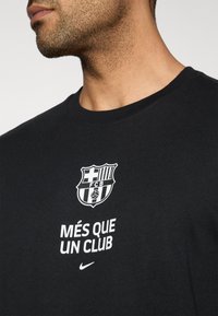 Μαύρο βαμβακερό μπλουζάκι με το έμβλημα "FCB" και το κείμενο "MÉS QUE UN CLUB" σε λευκό. Στρογγυλή λαιμόκοψη· κοντά μανίκια. Λογότυπο της Nike στο κάτω μέρος.