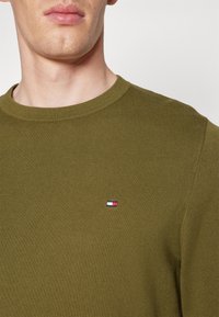 Homme portant un pull en tricot vert olive avec un petit logo rectangulaire rouge, blanc et bleu sur le côté gauche de la poitrine, sur un fond uni.