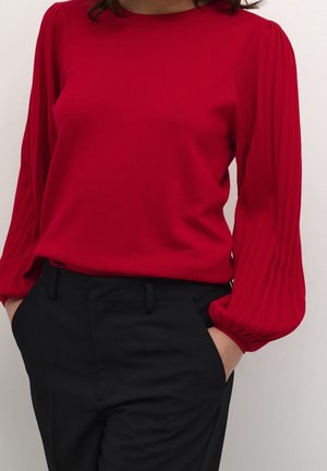 Donna che indossa una camicetta rossa a maniche lunghe con maniche plissettate e pantaloni neri, con le mani nelle tasche, su sfondo semplice.