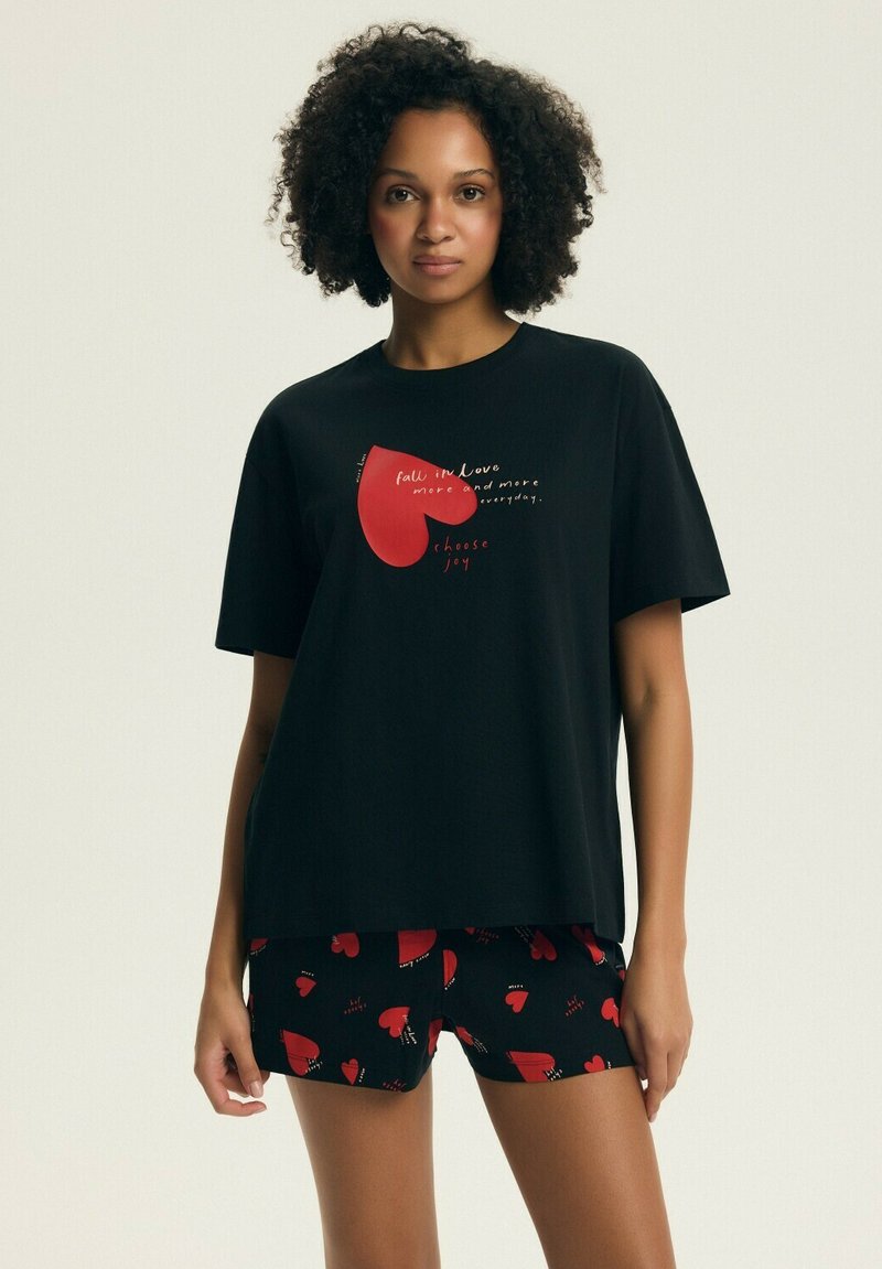 Kobieta z kręconymi włosami, ubrana w czarny zestaw t-shirt i szorty z czerwonymi wzorami serc oraz napisem o miłości i radości.