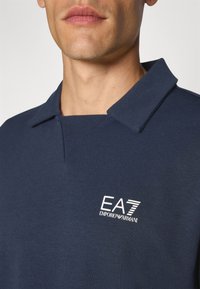 Temno modra polo majica z mehko teksturo, kratkim ovratnikom in belim izvezenim logotipom z napisom "EA7 EMPORIO ARMANI" na levi strani prsi.