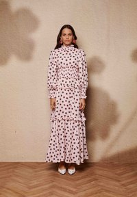 Μακριά ροζ φόρεμα με πουά, βολάν σε στρώσεις, μακριά μανίκια και κορσέ με σφηκοφωλιά. Συνδυασμένο με λευκά παπούτσια μυτερά με μαύρες βούλες.