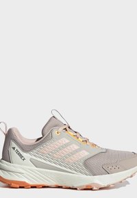 Ανοιχτό γκρι και μπεζ παπούτσι Adidas Terrex για τρέξιμο σε μονοπάτια με ροζ λεπτομέρειες και πορτοκαλί σόλα, εμφανίζεται από το πλάι σε λευκό φόντο.