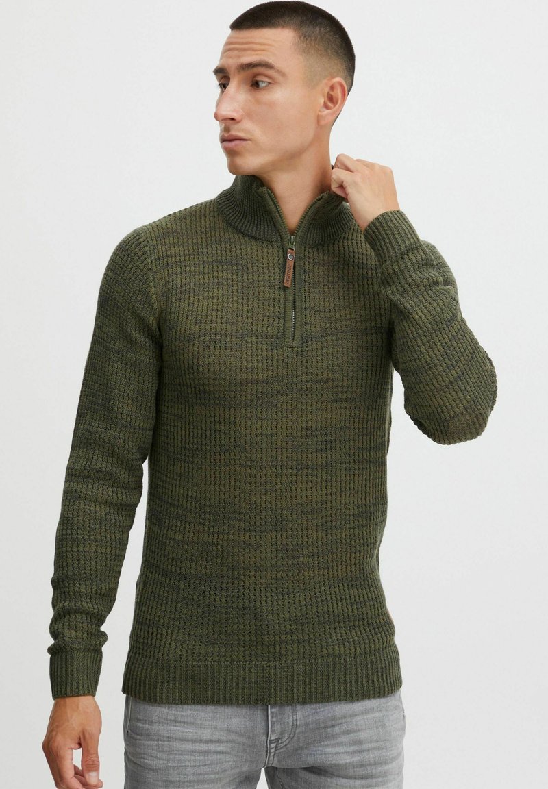 Grüner Strickpullover mit halbem Reißverschluss-Kragen, geripptem Material und umgeschlagenen Ärmeln. Mit einem Holz-Zipper für zusätzliche Details.