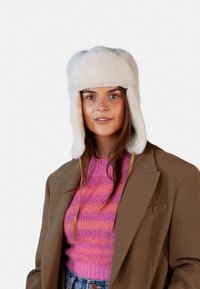 Gorro de piel blanca con solapas para las orejas, que presenta una textura suave y una tela resistente. Se lleva con un suéter a rayas en rosa y morado.