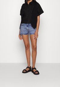 Osoba oblečená v černém tričku s krátkým rukávem, černé pletené vestě, modrých džínových šortkách a černých sandálech na pásku stojící na jednolitém pozadí.