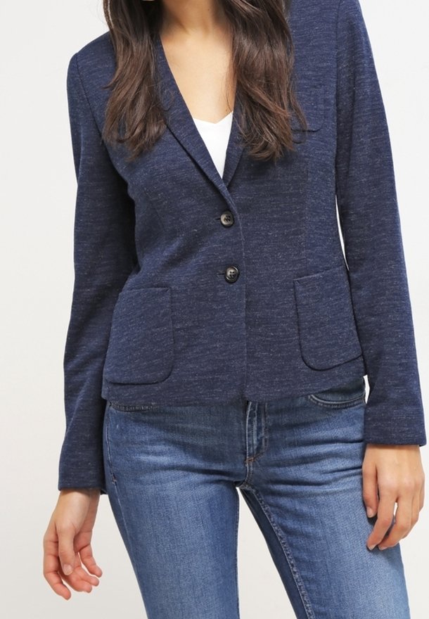 Femme portant un blazer bleu marine ajusté avec deux poches avant et un jean bleu foncé, debout avec une main détendue sur le côté.