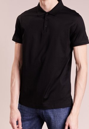 Mand iført en ensfarvet sort kortærmet poloshirt med tre knapper, kombineret med blå jeans, stående mod en neutral baggrund.