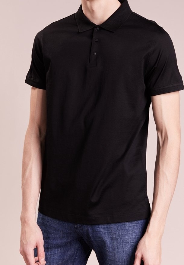 Homme portant un polo noir uni à manches courtes avec trois boutons, assorti à un jean bleu, debout devant un fond neutre.