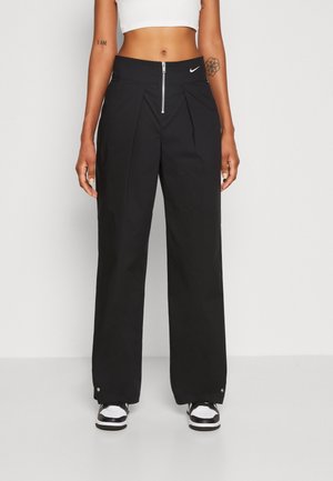 Frau trägt schwarze, weit geschnittene Hose mit Frontreißverschluss und Nike-Logo, weißes Croptop und schwarz-weiße Sneaker, steht vor einfarbigem Hintergrund.