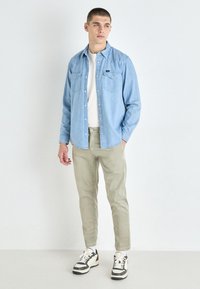 Lys blå denimskjorte, hvit t-skjorte med rund hals, olivengrønne bukser, svart og hvite sneakers. To brystlommer på skjorten.