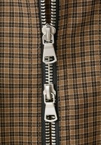 Prim-plan al unui material maro în carouri cu un fermoar negru cu două capete argintii, unul situat deasupra celuilalt.