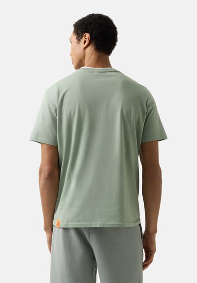 Jeune homme vu de dos portant un t-shirt vert clair uni à manches courtes et un pantalon gris clair sur fond blanc.