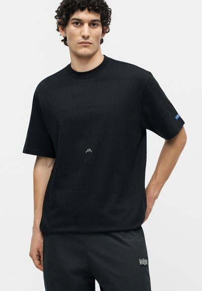 Νέος άνδρας με σγουρά μαλλιά που φοράει μια απλή μαύρη φαρδιά μπλούζα και μαύρο παντελόνι, στέκεται με το ένα χέρι στη μέση σε λευκό φόντο.