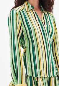 Blouse rayée vert, jaune et noir. Présente un col avec un encolure et une texture douce. Manches longues avec une coupe légèrement décontractée.